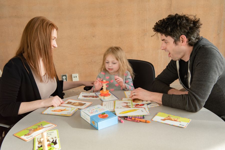 A four-year-old girl sits with her parents (mom, left; dad, right) with educational tools in front of her.