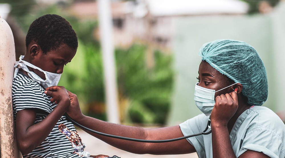 An African doctor wearing a facemask and hairnet holds a stethoscope up to a child's chest.