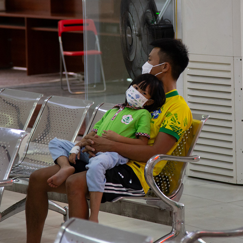 A man sits on a chair, holding a barefoot child, both wearing facemasks