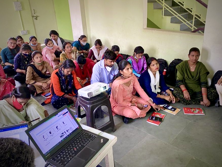 A group of male and female healthcare providers in India, with medium light to medium dark skin tones, sit on a floor and look towards a presentation during an ECHO session.