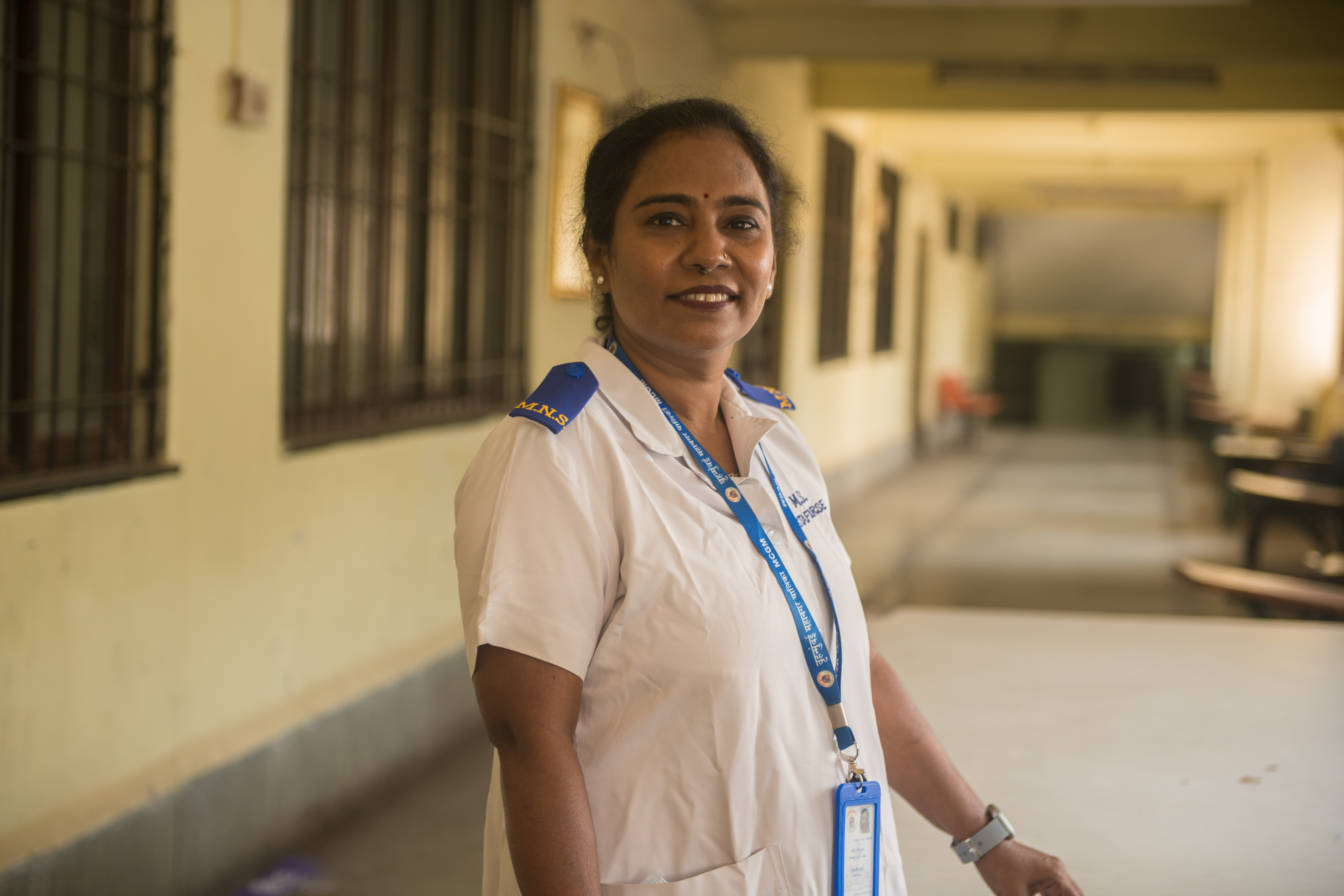 A nurse in India wearing a white uniform and a badge stands confidently and smiles.