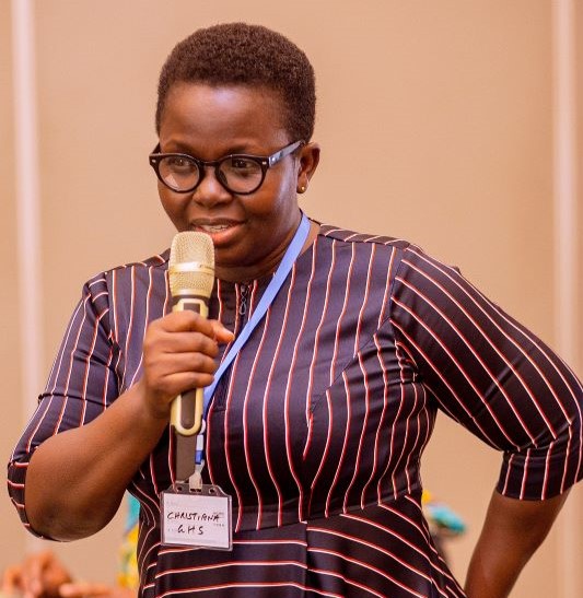A female presenter, standing, speaks at a conference; photo taken in Ghana.