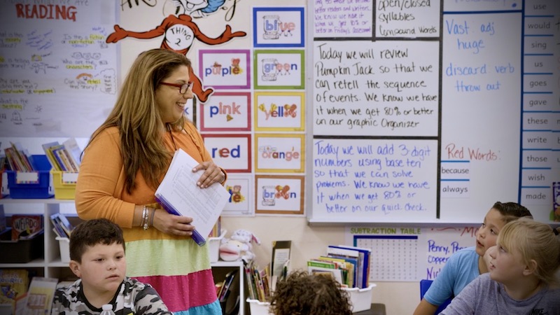 A female teacher stands in a classroom smiling at her students, two children look at her