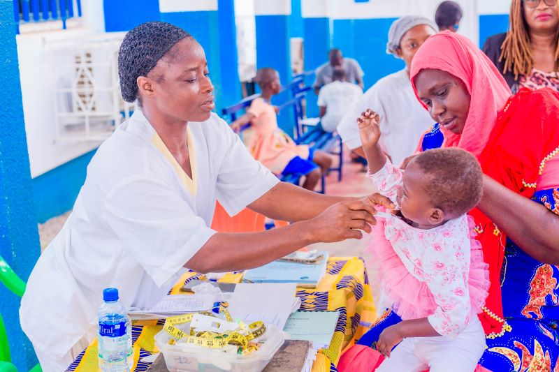 A female African health care provider reaches across a table to a young girl sitting on her mother's lap.