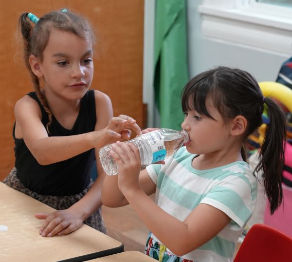 Two young girls sitting at a table; one helps the other drink from a bottle of water