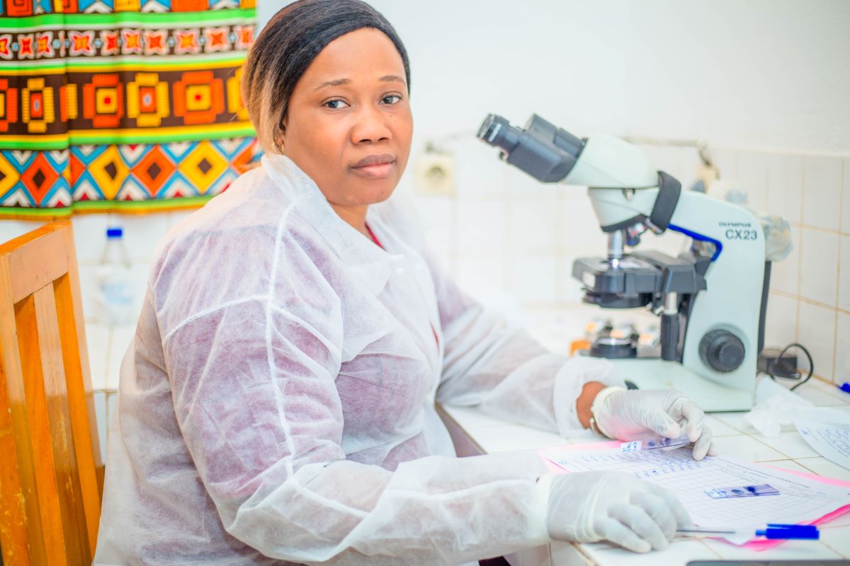 An African woman in a laboratory, wearing protective gear. There is a microscope in the background