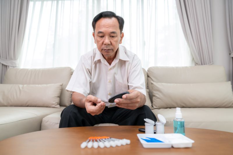 A stock image: Asian man sits in a living room with his glucose monitoring equipment in front of him. He pricks his finger