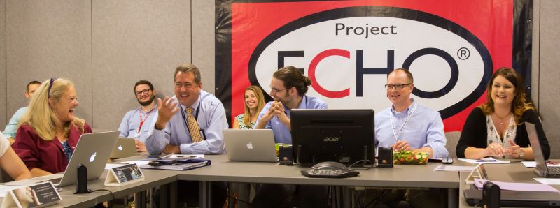 A group of professionally dressed people, with laptops in front of them, sit at a conference table. They all wear smiles, reacting to one person speaking.