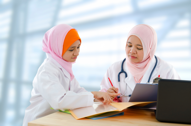 A stock image of two women, wearing head coverings and white lab coats, look together at a piece of paper.