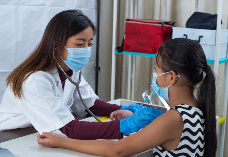 A female health care worker holds a stethoscope to listen to a young female child's heart. Both wear face masks.