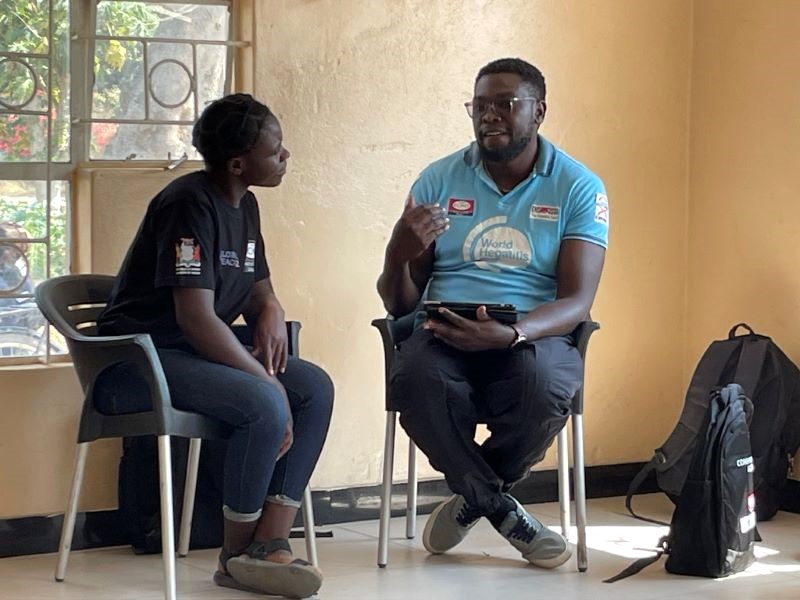 Taken in Zambia, a man, right, wears a World Health Organization blue t-shirt and speaks with someone on the left. Both are seated.