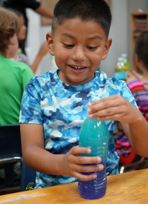 A Latino boy, about five years old, holds the cap on a water bottle. The water has been dyed blue in preparation of a science experiment.