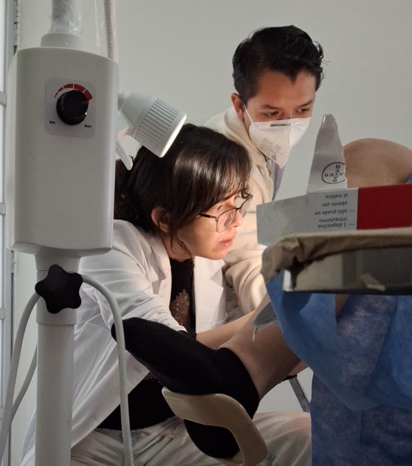 A young female doctor looks between a patient's legs (feet and gown visible). A male clinician, wearing a face mask, looks from beside her.