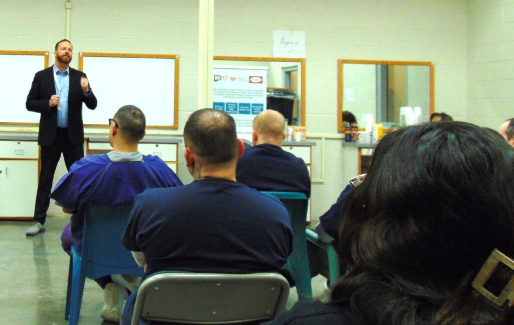 A man in a suit stand at the front of a room wearing a suit. Incarcerate people, wearing dark blue, sit listening to him