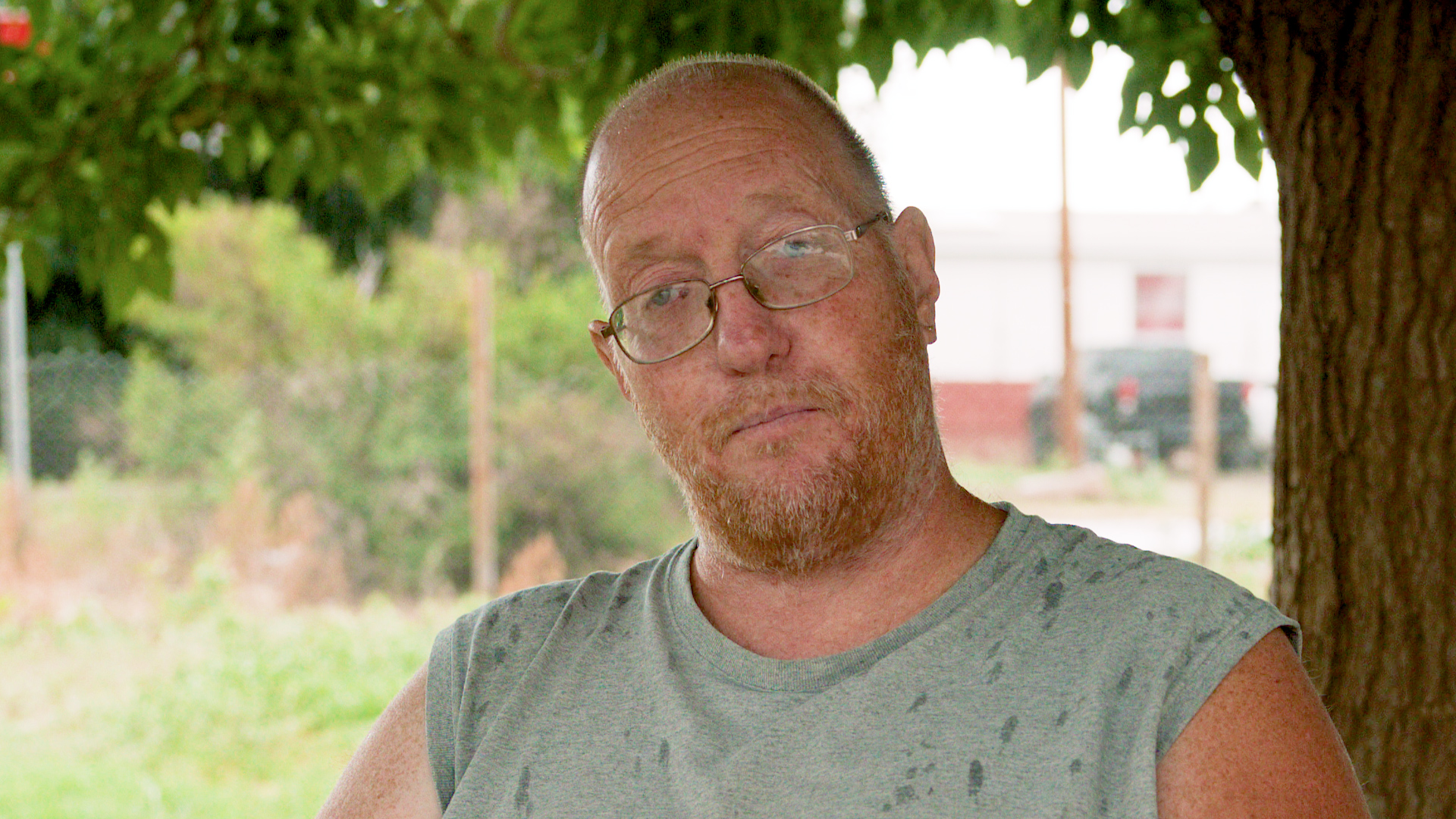 Tim Loflin, of Clovis, NM, is an older white male. His sits outside, underneath a tree with rain drops on his shirt.