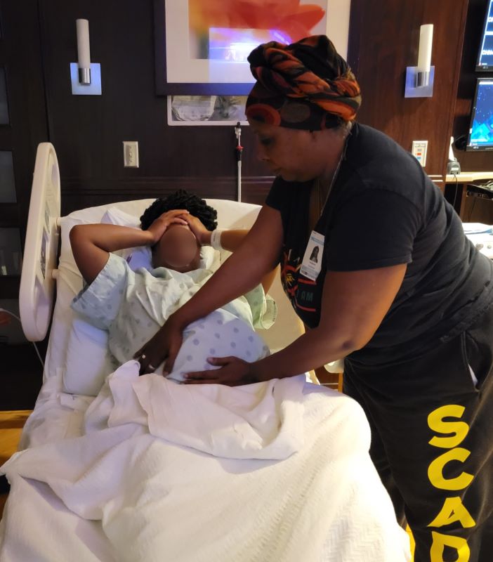 A Black woman places her hands on a pregnant woman's belly. They are in a hospital room.