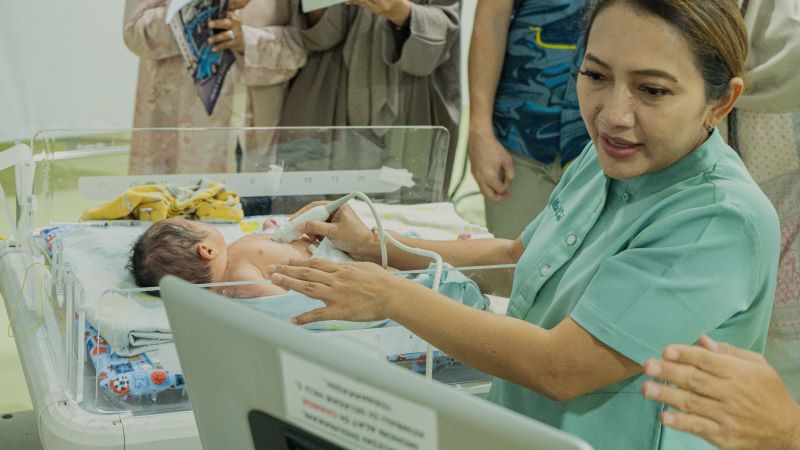 A female provider, wearing green scrubs, holds a medical device to a newborn's heart and looks at a monitor.