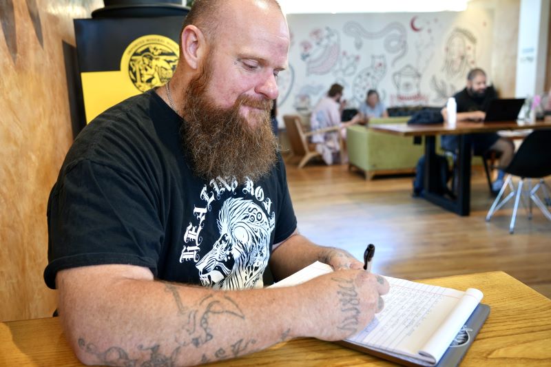 Matthew Pettite, white male, sits in a coffeeshop in Albuquerque, NM. He writes on a notepad.