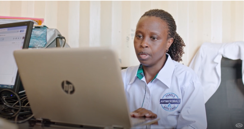 A female provider looks at a laptop in front of her. Her shirt reads "Handle Antimicrobials with Care."