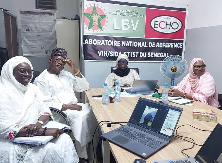 Four Senegalese people (three women, one man), wearing traditional clothing and head coverings, sit around a table. A laptop with a Zoom meeting is also in the picture.