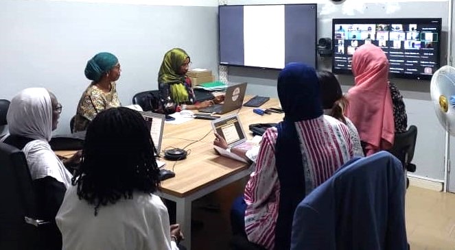 Six Senegalese women, five wearing headscarves, sit at a conference table and look at a monitor