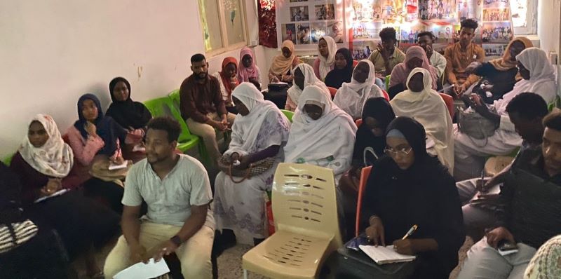 Men and women sit in a classroom and look ahead. Some take notes. The women war head coverings and traditional clothes.