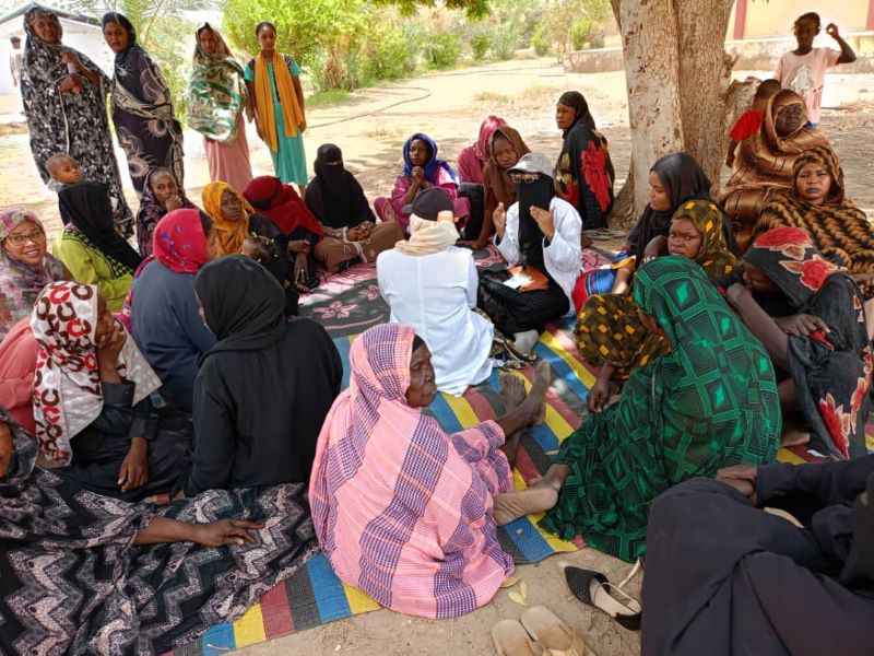 A group of women, all wearing traditional clothing and headscarves, sit on blankets in the shade. They listen to a masked woman in a white coat.