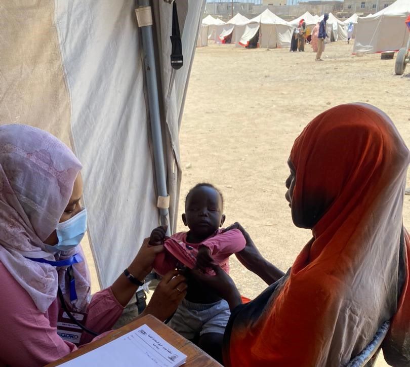 Two women, one wearing a mask and a stethoscope, hold a baby between them. Both women wear head coverings.