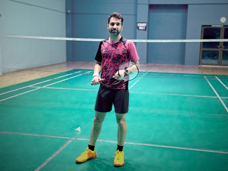Kartik Dhar holds a badminton raquet and stands on a badminton court.
