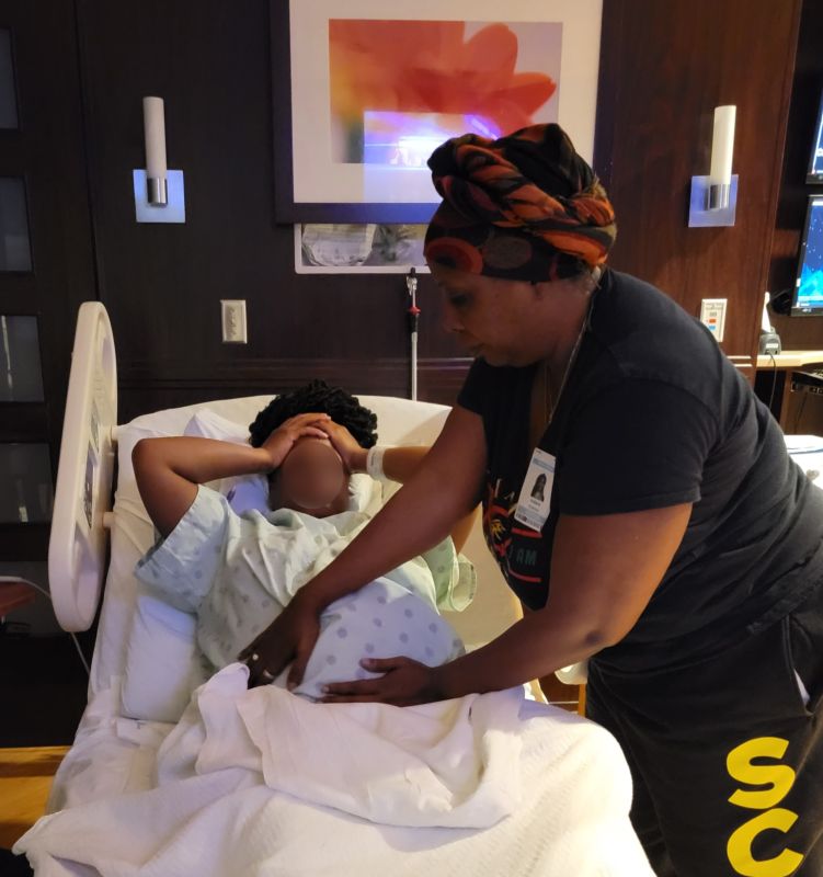 A Black woman, with a blurred face for privacy, is in a hospital bed giving birth. Her doula stands above and feels her stomach.