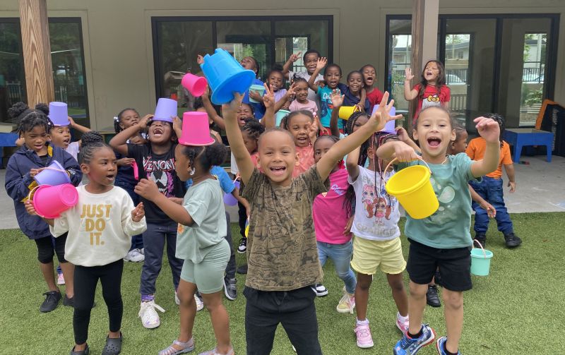A group of young children hold up colorful bucket and shout toward the camera