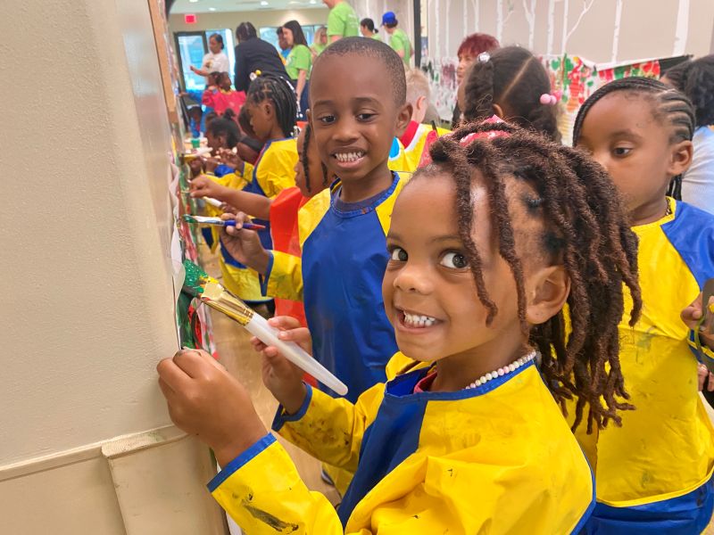 Elementary aged Black children wear smocks and do art on a wall. A young care (front, center) smiles and looks at the camera.