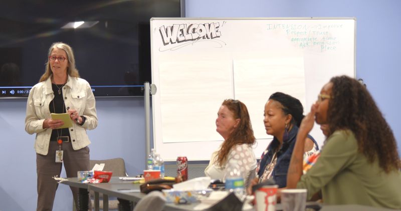 Dr. Karla Thornton (left) stands at the front of a room. Three women, seated, pay attention to something on the other side of the room.