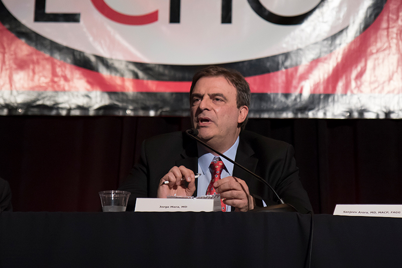 Dr. Jorge Mera sits at a conference table, speaking into a microphone during a presentation