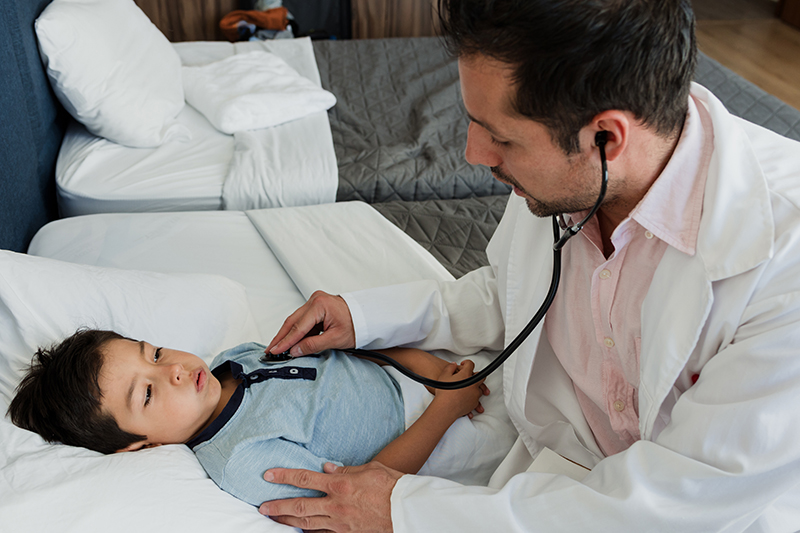 A doctor using a stethoscope to listen to a young patient's heart in a hospital bed. The patient is lying down in a hospital setting.