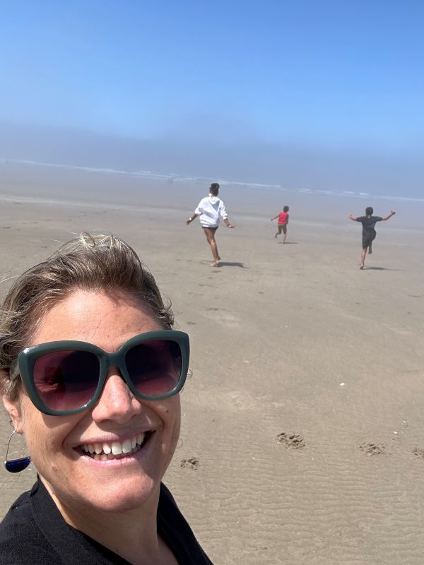 Simran Priel takes a selfie on a beach. In the background, three kids run toward the water.