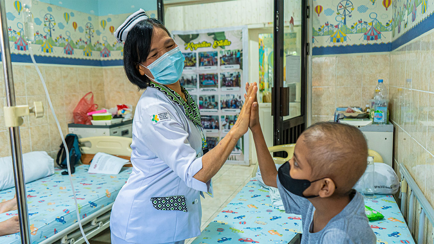 A nurse wearing a mask high-fives a child in a mask sitting on a hospital bed.