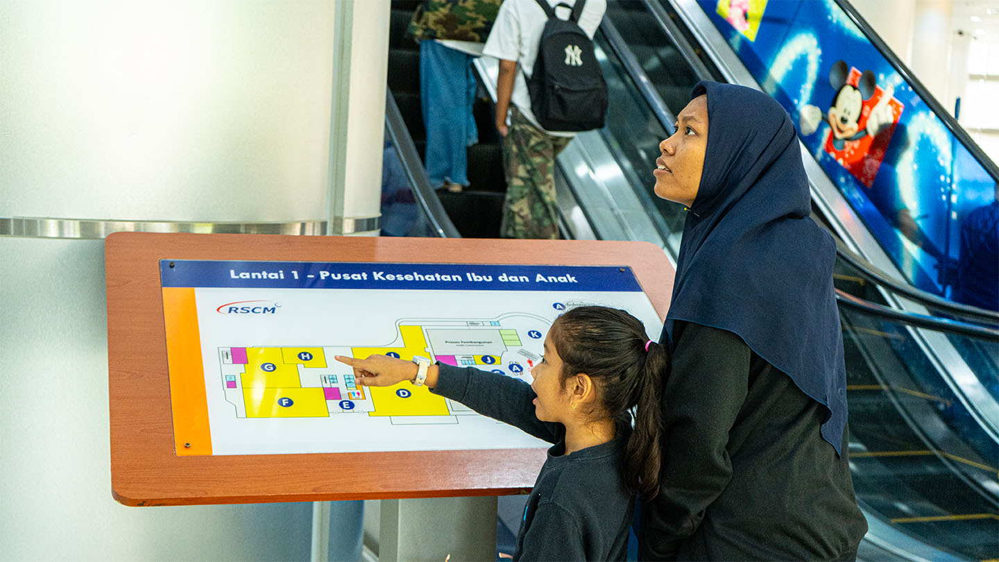 A mother in a head scarf with her daughter in front of a map kiosk in an Indonesian hospital with escalators visible in the background.