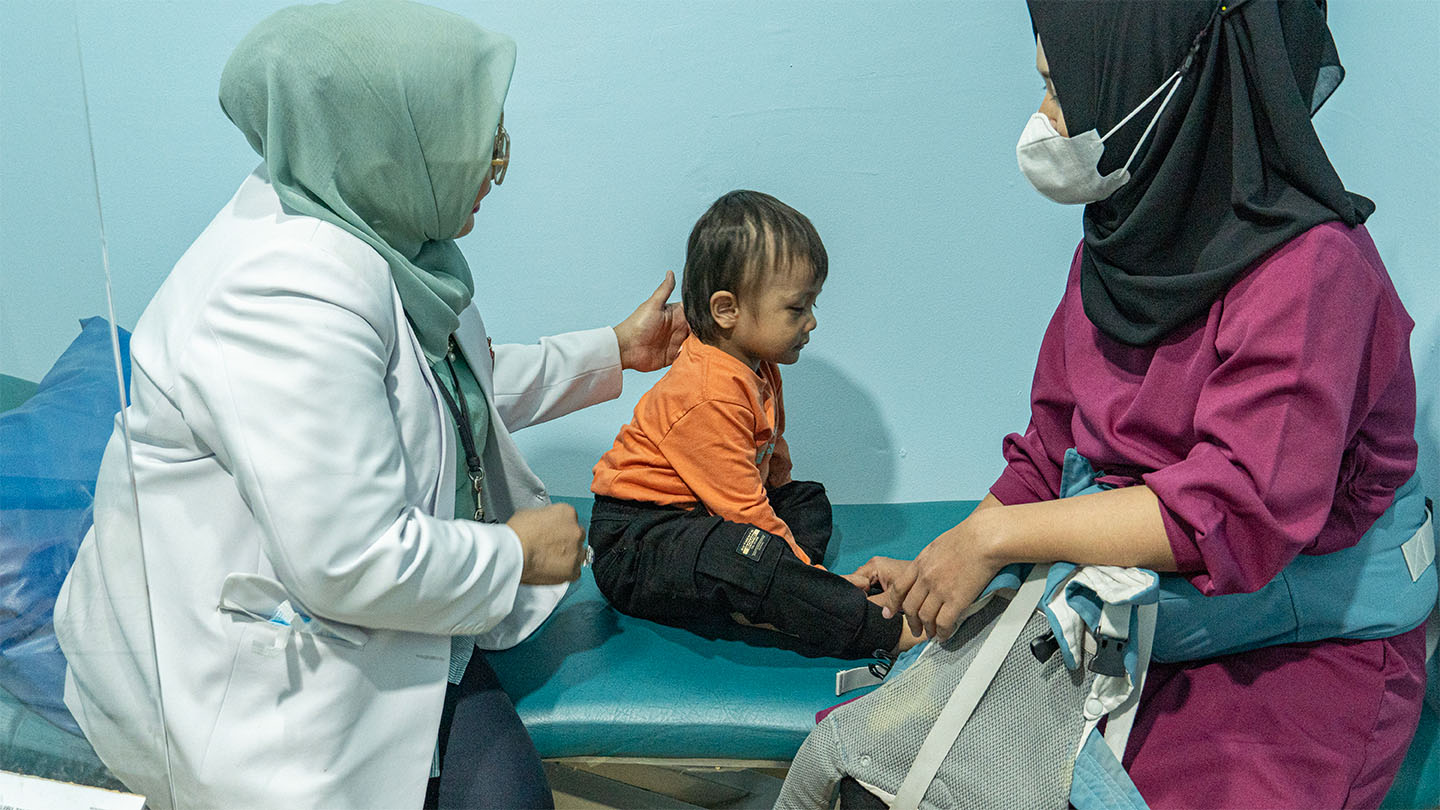 A woman in a white coat and head scarf gently touches a male toddler's back. Across from them, a woman in a mask and head scarf looks at the boy.
