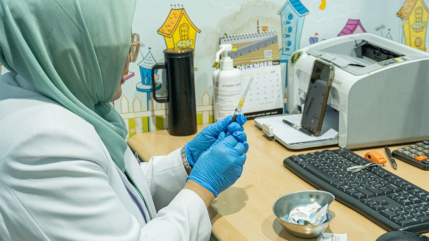 A nurse in Indonesia wearing a head scarf and gloves preparing a syringe.