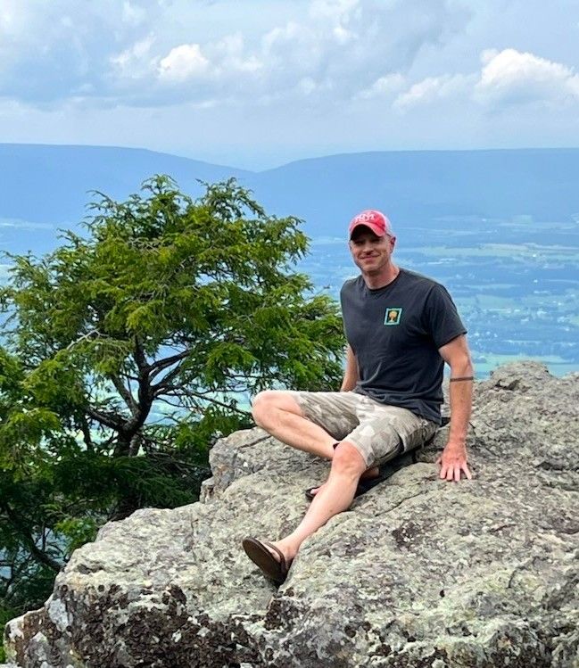 Dr. Matthew Bouchonville sits on top of a rock. You can see a valley and mountains behind him.