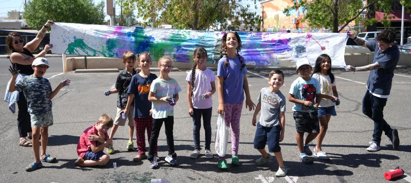 A group of young children stand in front of a cloth they have tie-dyed. Their teachers hold it behind them.