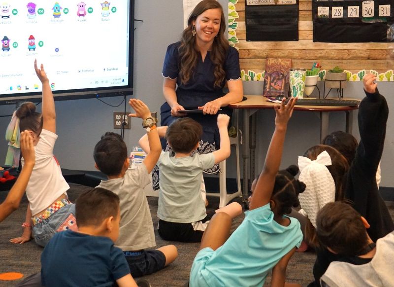 A young Caucasian teacher sits at the front of her classroom. Elementary-aged children raise their hands.