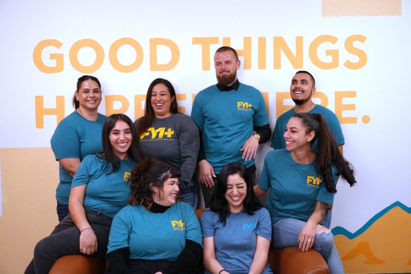 A group of eight people, wearing FYI+ t-shirts, sit and stand together.