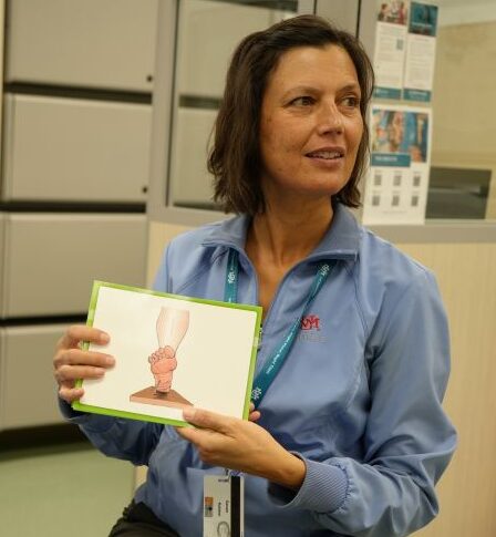 A woman, wearing a blue UNM Hospitals shirt and badge, holds up a drawing of a foot pointed upwards.