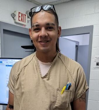 An incarcerated person, wearing beige, stands in front of a popsicle tower that has two awards on it.
