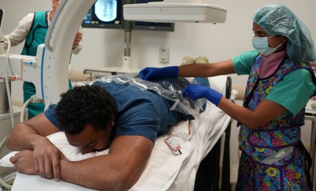 A male patient lies faces down under a machine. A health care provider, wearing PPE, works on his back.