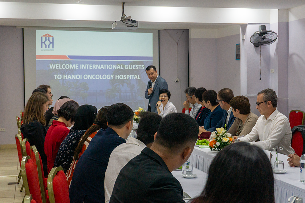A conference room filled with professional. A slide read "Welcome International Guests to Hanoi Oncology Hospital"