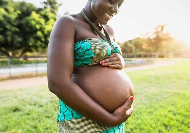 An African woman, wearing a teal bikini, holds her pregnant belly, smiling