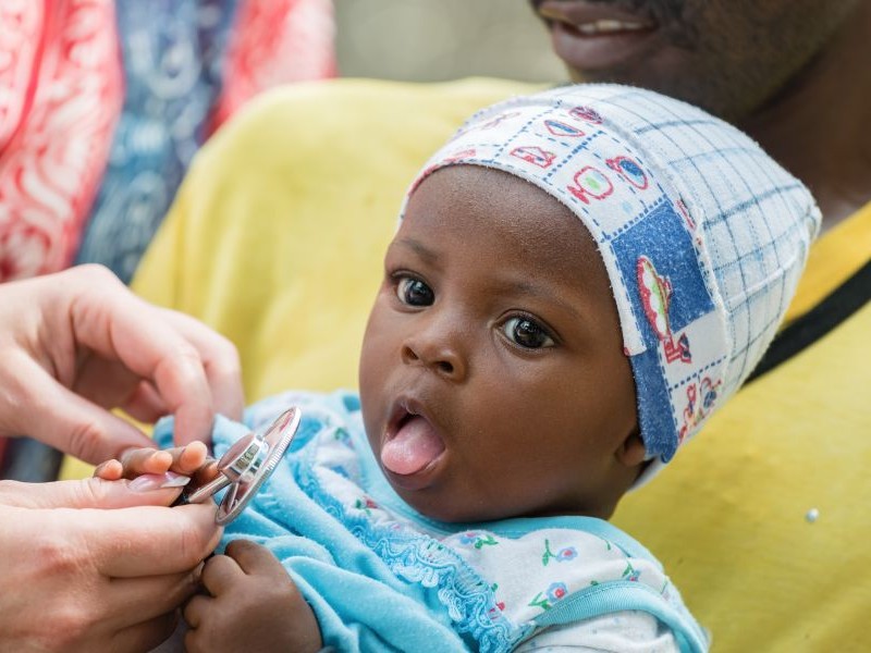 An infant with their tongue sticking out. A health care provider holds a stethoscope near them.